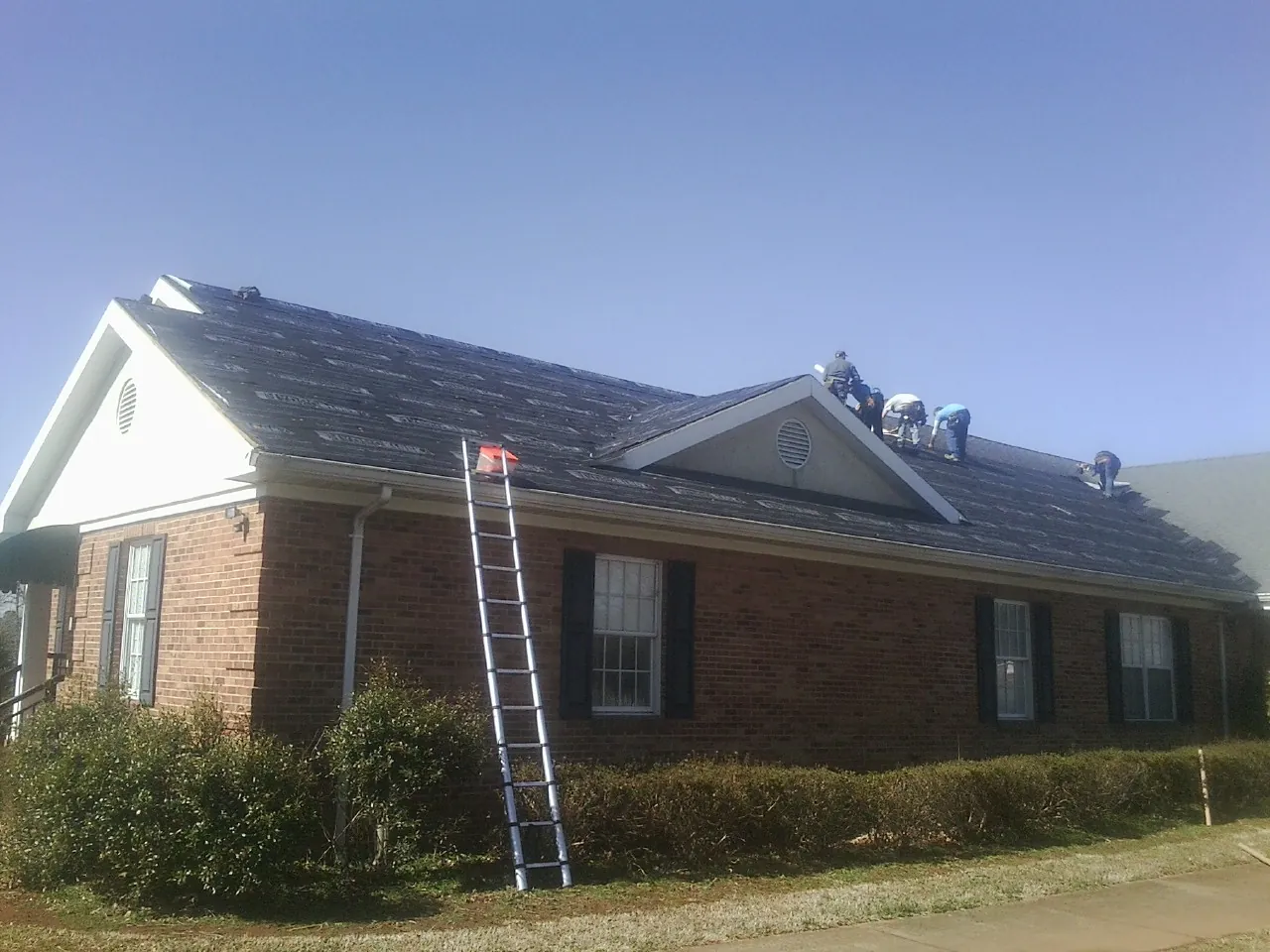 Crew tearing shingles off a brick ranch roof with a ladder set — Emergency Roof Repair work by our crew in Ada